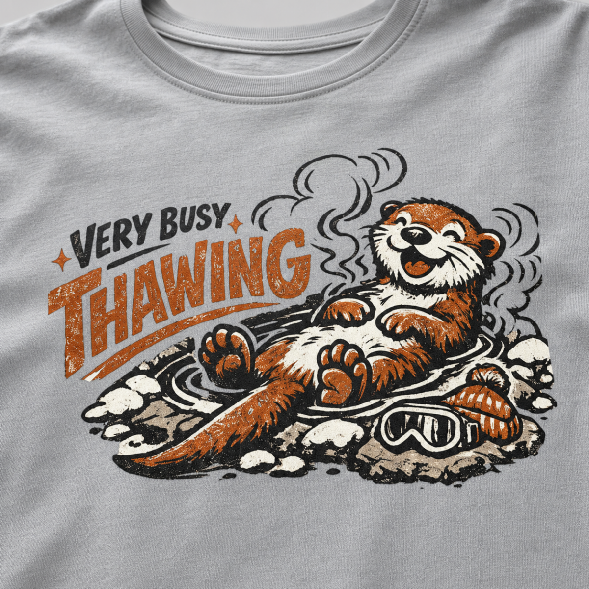 Close-up of a granite gray t-shirt featuring a relaxed otter soaking in a steaming hot spring beneath “Very Busy Thawing” text.
