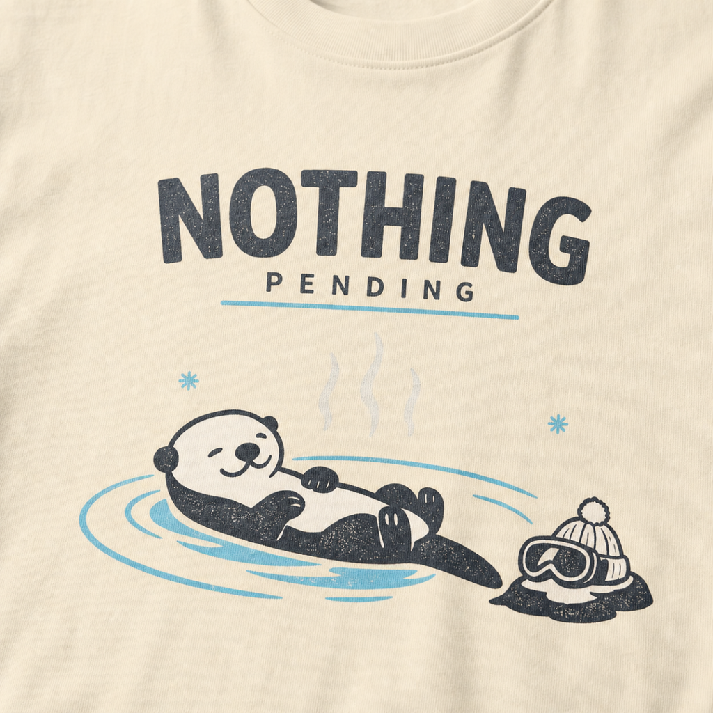 Close-up of an ivory white t-shirt featuring a relaxed otter floating in a hot spring beside ski goggles and a beanie, with the words “Nothing Pending.”