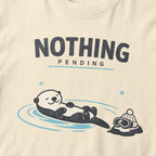 Close-up of an ivory white t-shirt featuring a relaxed otter floating in a hot spring beside ski goggles and a beanie, with the words “Nothing Pending.”