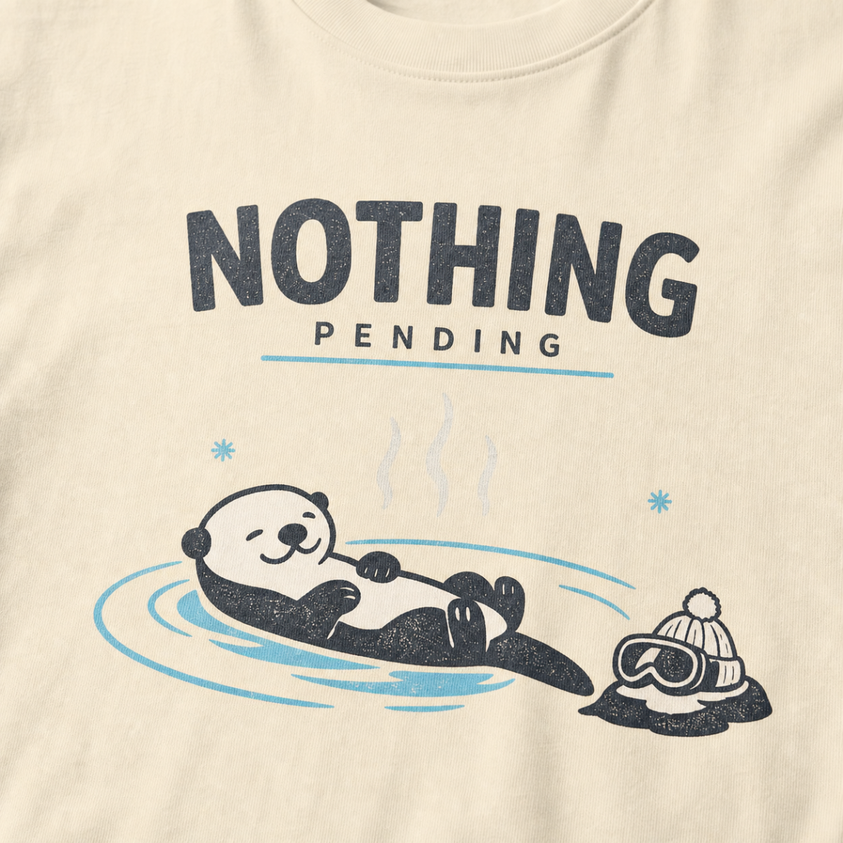 Close-up of an ivory white t-shirt featuring a relaxed otter floating in a hot spring beside ski goggles and a beanie, with the words “Nothing Pending.”