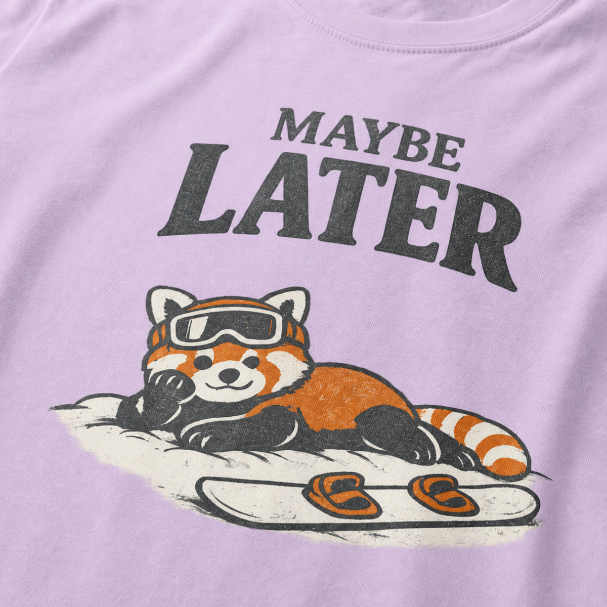 Close-up of an orchid purple t-shirt design featuring a red panda resting in the snow beside a snowboard beneath the words “Maybe Later.”
