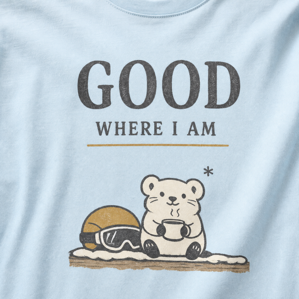 Close-up of a chambray blue t-shirt featuring a small white bear holding a warm drink beside ski goggles with the phrase “Good Where I Am.”
