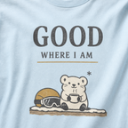 Close-up of a chambray blue t-shirt featuring a small white bear holding a warm drink beside ski goggles with the phrase “Good Where I Am.”