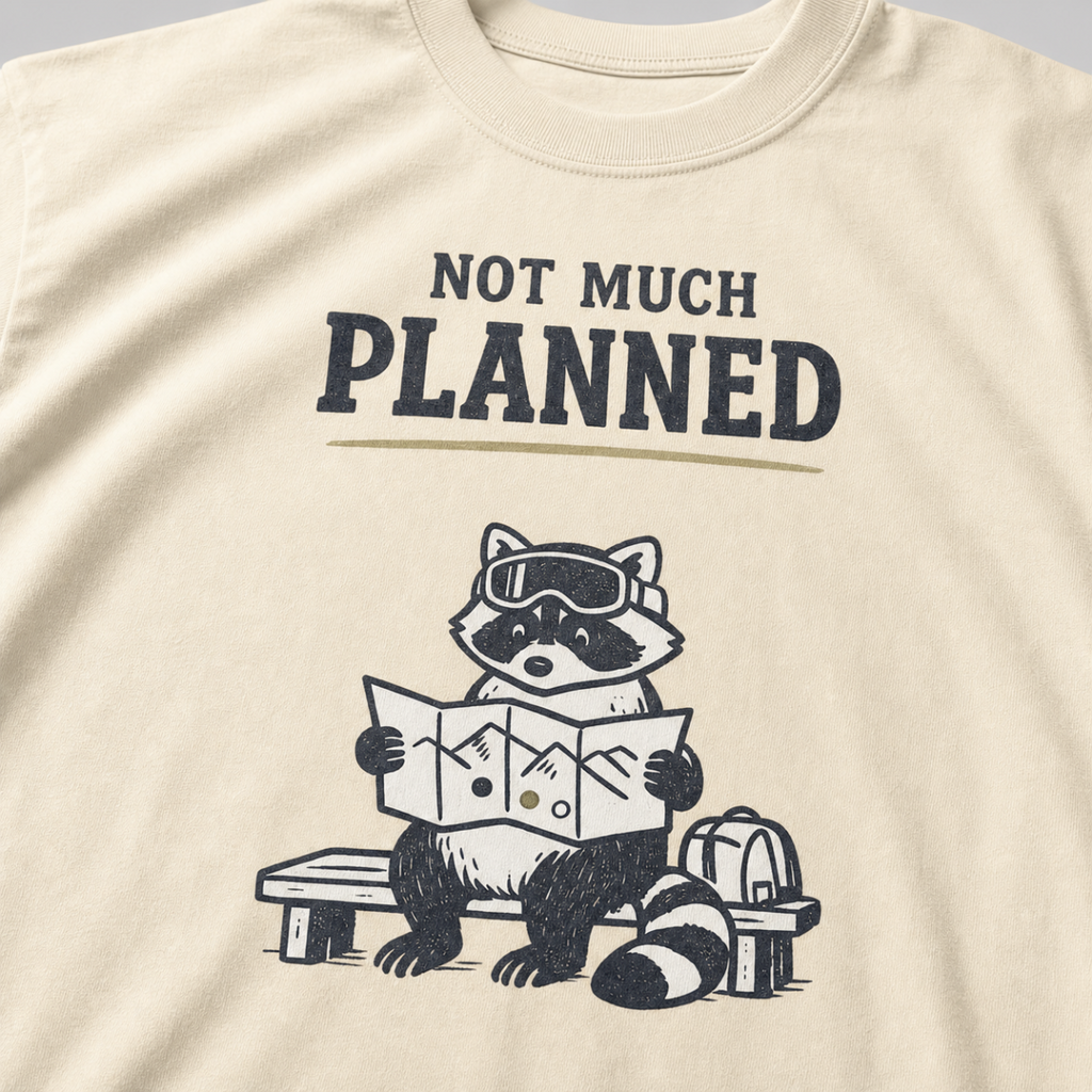 Close-up of an ivory white t-shirt featuring a raccoon in ski goggles holding a mountain map beside a backpack, with the words “Not Much Planned.”