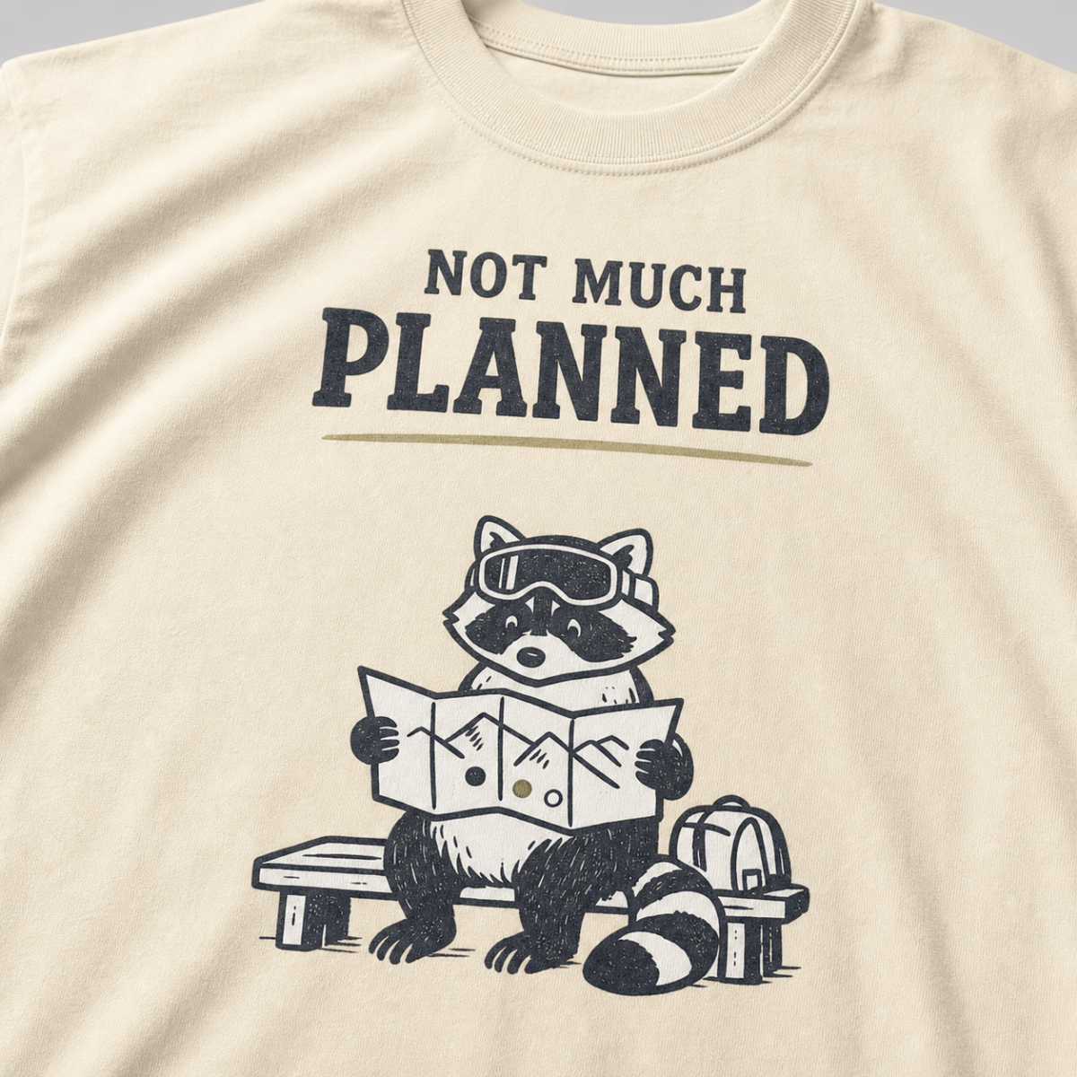 Close-up of an ivory white t-shirt featuring a raccoon in ski goggles holding a mountain map beside a backpack, with the words “Not Much Planned.”