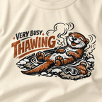 Close-up of an ivory white t-shirt featuring a relaxed otter soaking in a steaming hot spring beneath “Very Busy Thawing” text.