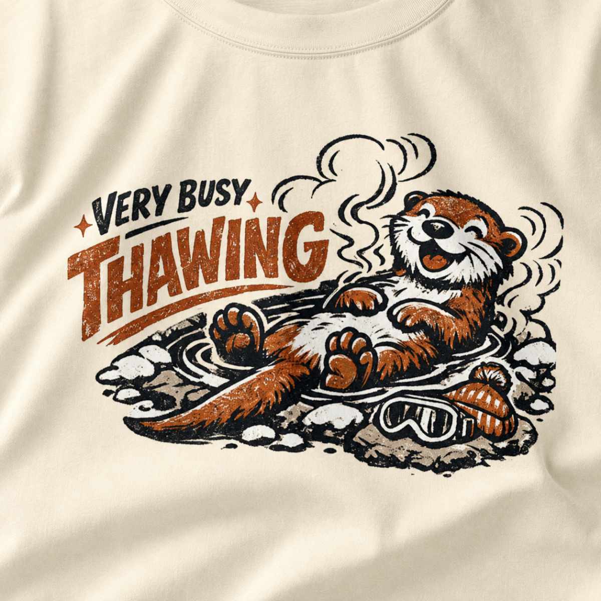 Close-up of an ivory white t-shirt featuring a relaxed otter soaking in a steaming hot spring beneath “Very Busy Thawing” text.