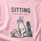 Close-up of a rabbit in ski goggles sitting beside a mug and snowboard beneath “Sitting This One Out” lettering.