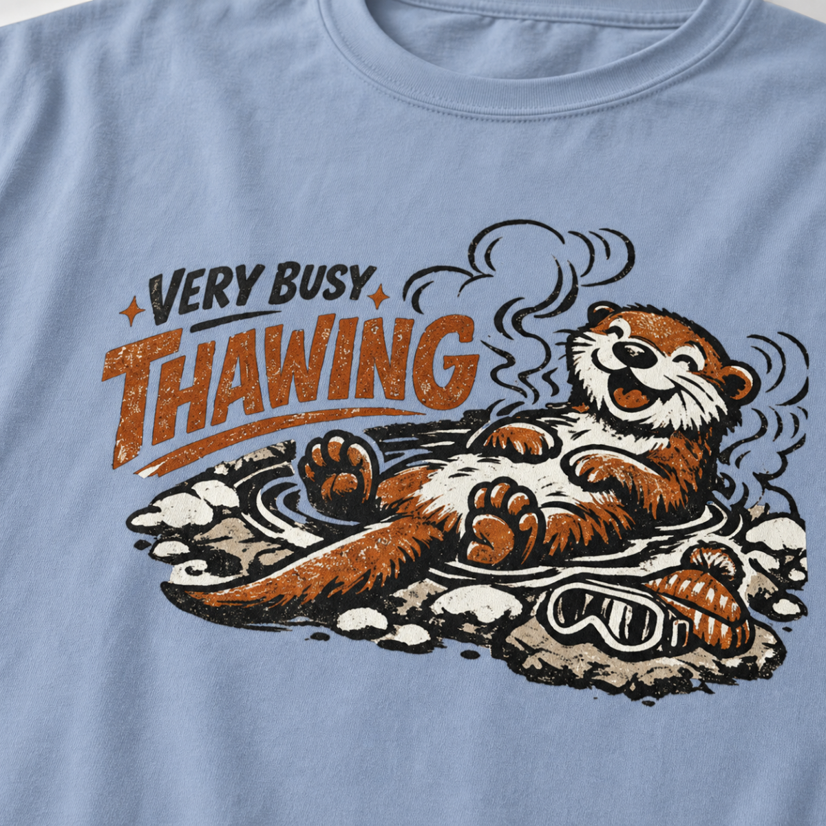 Close-up of a washed denim blue t-shirt featuring a relaxed otter soaking in a steaming hot spring beneath “Very Busy Thawing” text.