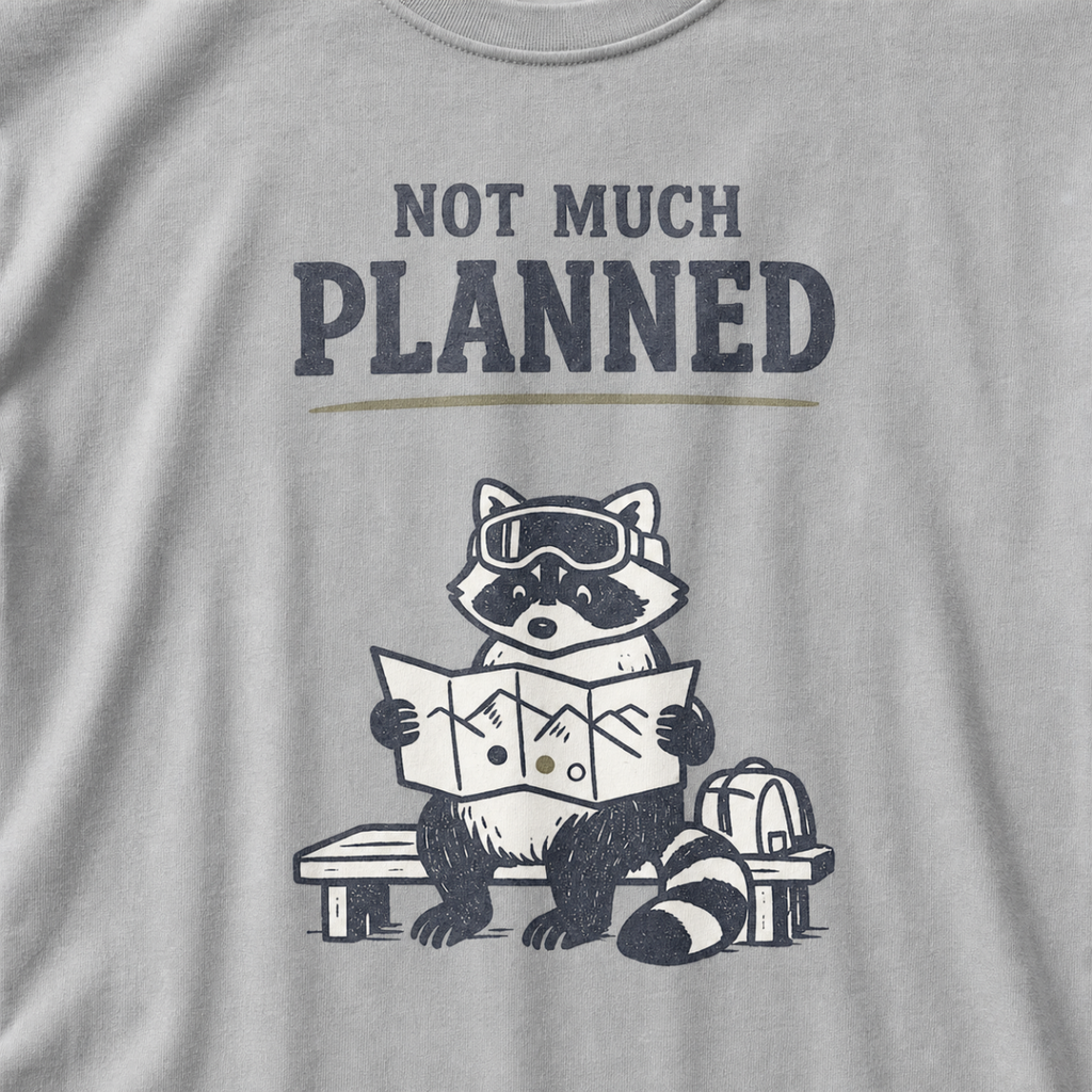 Close-up of a granite gray t-shirt featuring a raccoon in ski goggles holding a mountain map beside a backpack, with the words “Not Much Planned.”