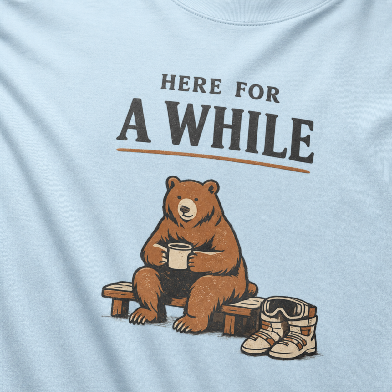 Close-up of a chambray blue t-shirt featuring a bear holding a mug beside ski boots and goggles with the phrase “Here for a While.”
