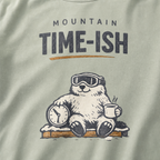 Close-up of a bay green t-shirt design showing a relaxed marmot with ski goggles, a clock and steaming mug beneath the words “Mountain Time-ish.”