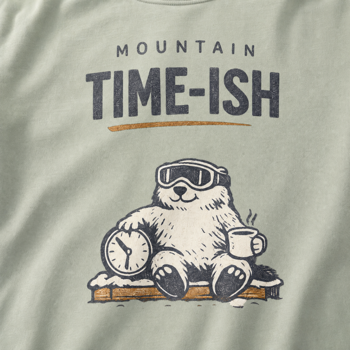 Close-up of a bay green t-shirt design showing a relaxed marmot with ski goggles, a clock and steaming mug beneath the words “Mountain Time-ish.”