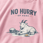 Close-up of a blossom pink t-shirt featuring a mountain goat resting in the snow beside a helmet, goggles and snack beneath the words “No Hurry Up Here.”