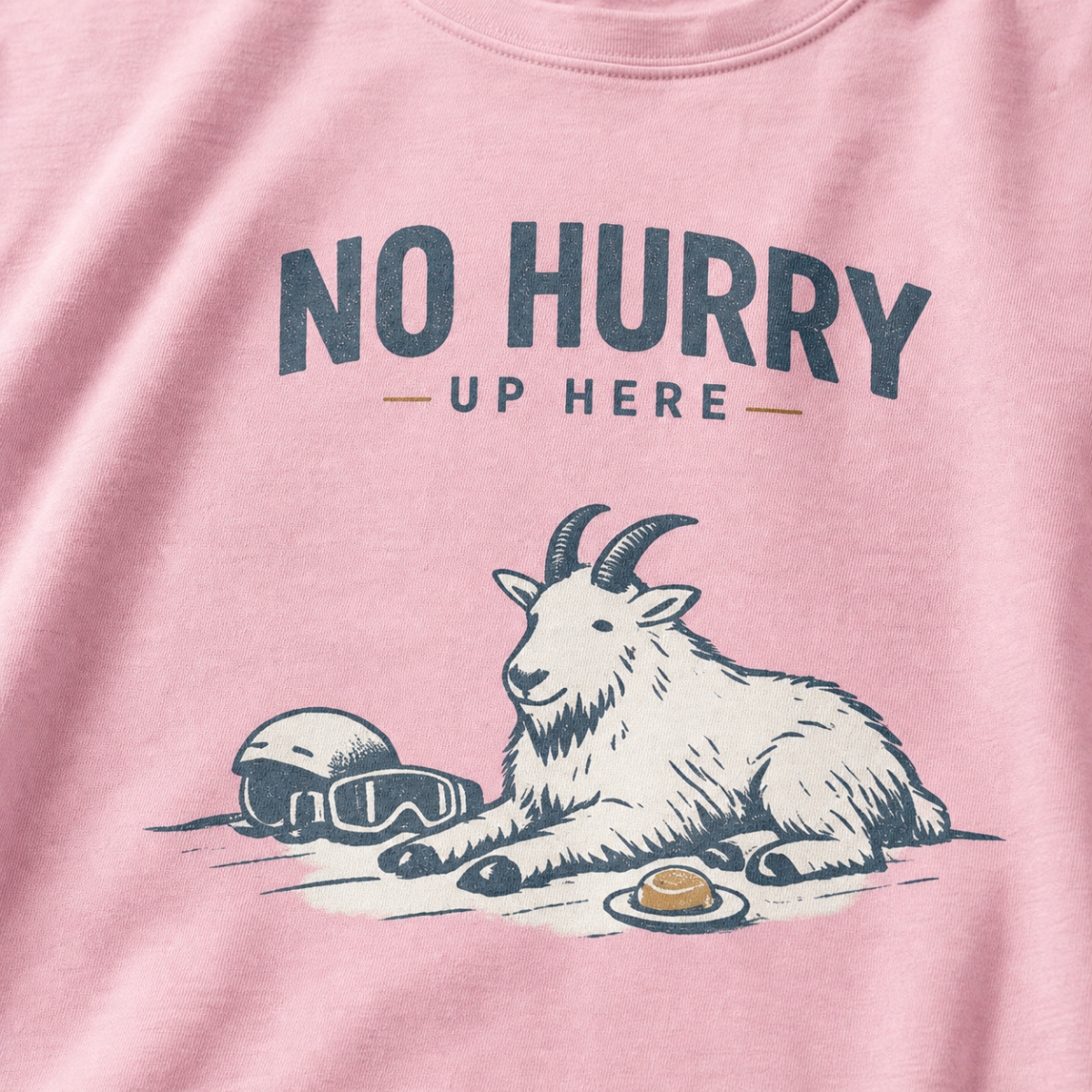 Close-up of a blossom pink t-shirt featuring a mountain goat resting in the snow beside a helmet, goggles and snack beneath the words “No Hurry Up Here.”