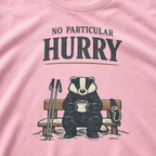 Close-up of a blossom pink t-shirt featuring a badger seated on a snowy bench with a mug, ski poles and goggles beneath the words “No Particular Hurry.”