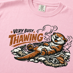 Close-up of a blossom pink t-shirt featuring a relaxed otter soaking in a steaming hot spring beneath “Very Busy Thawing” text.