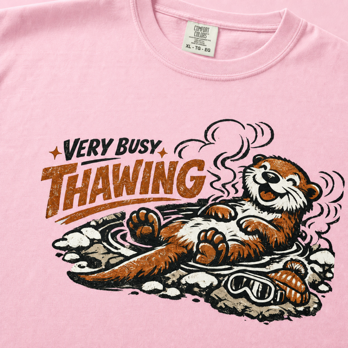 Close-up of a blossom pink t-shirt featuring a relaxed otter soaking in a steaming hot spring beneath “Very Busy Thawing” text.