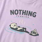 Close-up of an orchid purple t-shirt featuring a relaxed otter floating in a hot spring beside ski goggles and a beanie, with the words “Nothing Pending.”