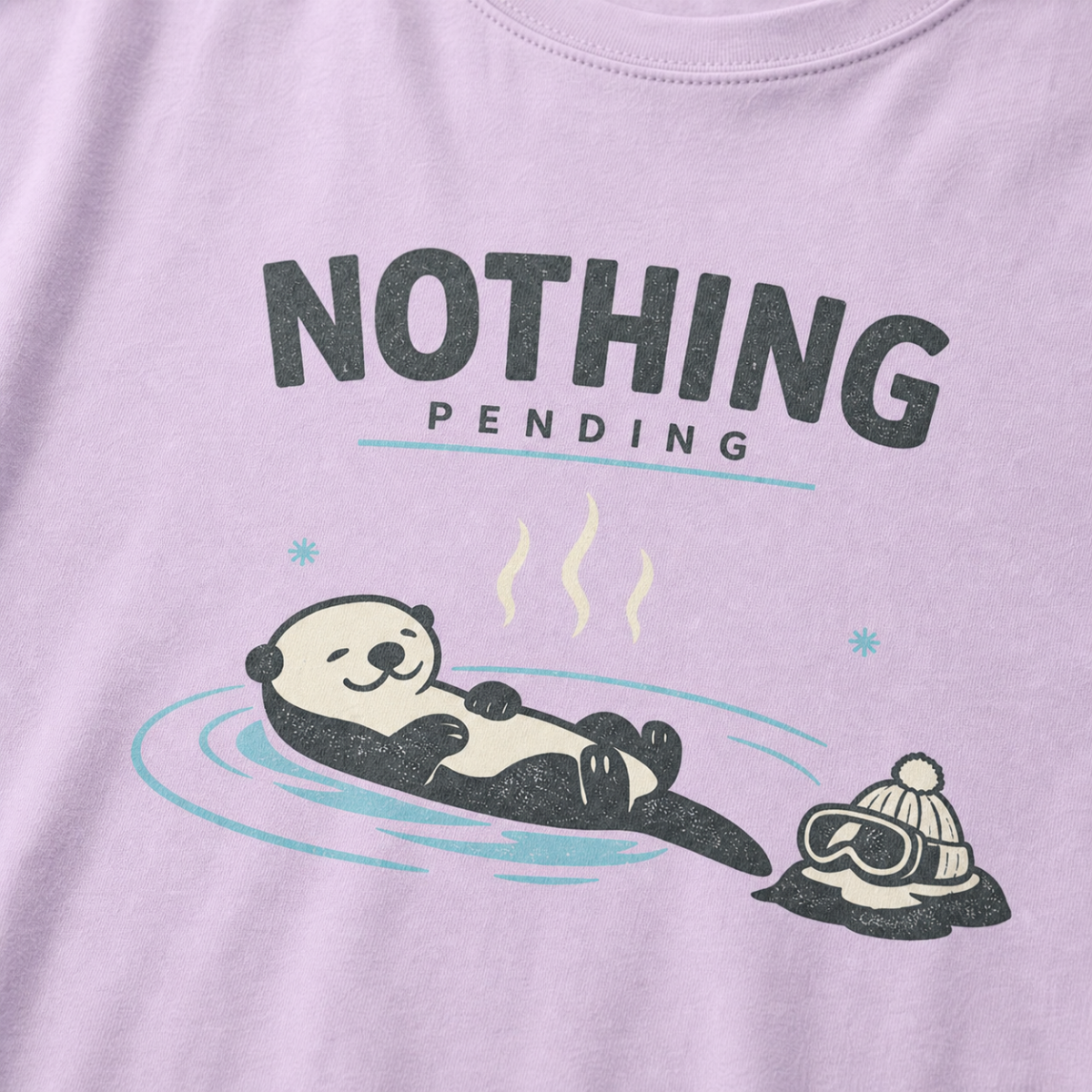 Close-up of an orchid purple t-shirt featuring a relaxed otter floating in a hot spring beside ski goggles and a beanie, with the words “Nothing Pending.”
