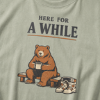 Close-up of a bay green t-shirt featuring a bear holding a mug beside ski boots and goggles with the phrase “Here for a While.”
