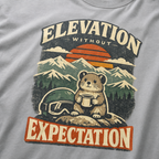 Close-up of a granite grey t-shirt design featuring a small mountain rodent holding a mug beside ski goggles, with sunset peaks and the phrase “Elevation Without Expectation.”