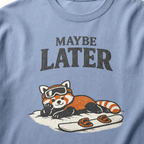Close-up of a washed denim blue t-shirt design featuring a red panda resting in the snow beside a snowboard beneath the words “Maybe Later.”
