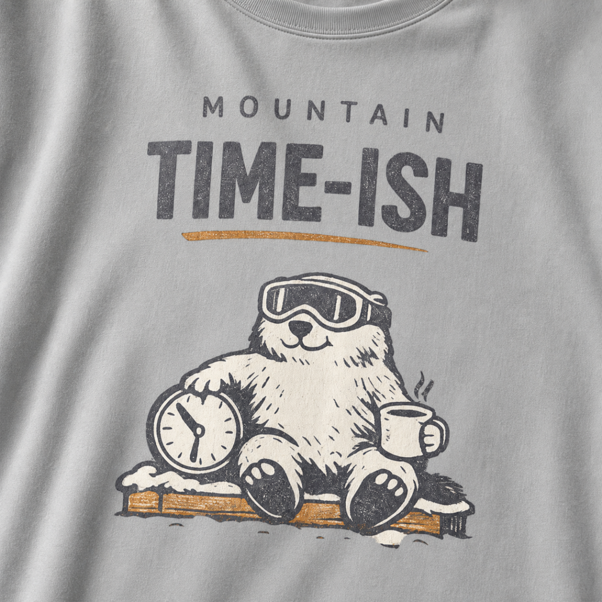 Close-up of a granite gray t-shirt design showing a relaxed marmot with ski goggles, a clock and steaming mug beneath the words “Mountain Time-ish.”