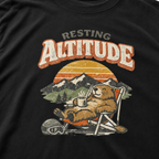 Close-up of a relaxed marmot reclining in a chair with a steaming mug beneath “Resting Altitude” lettering and a mountain sunset.