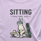Close-up of a rabbit in ski goggles sitting beside a mug and snowboard beneath “Sitting This One Out” lettering.