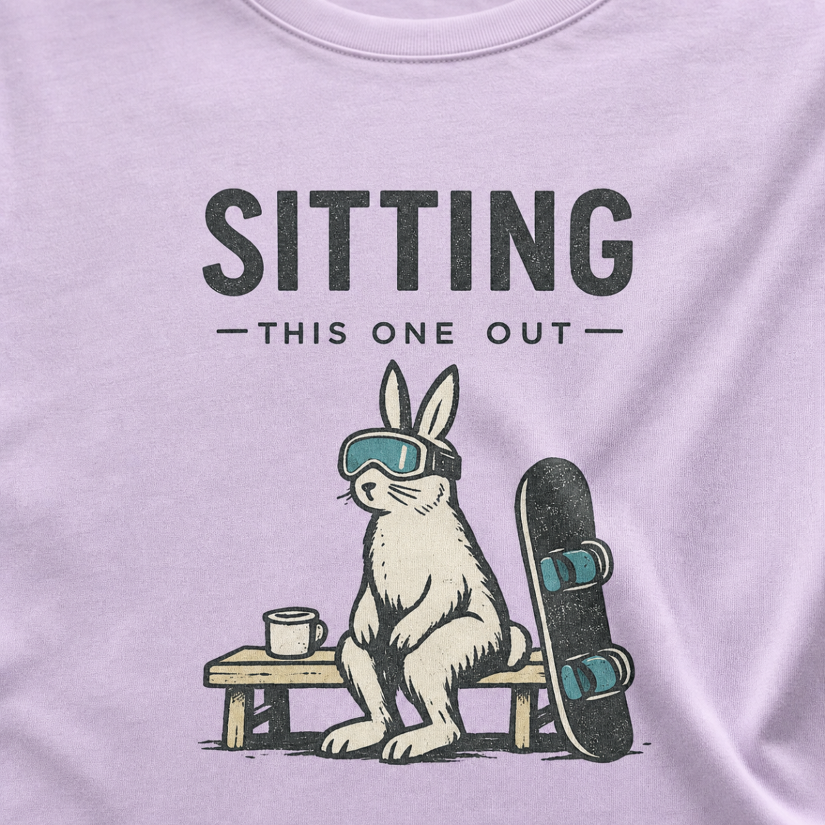 Close-up of a rabbit in ski goggles sitting beside a mug and snowboard beneath “Sitting This One Out” lettering.