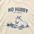 Close-up of an ivory white t-shirt featuring a mountain goat resting in the snow beside a helmet, goggles and snack beneath the words “No Hurry Up Here.”