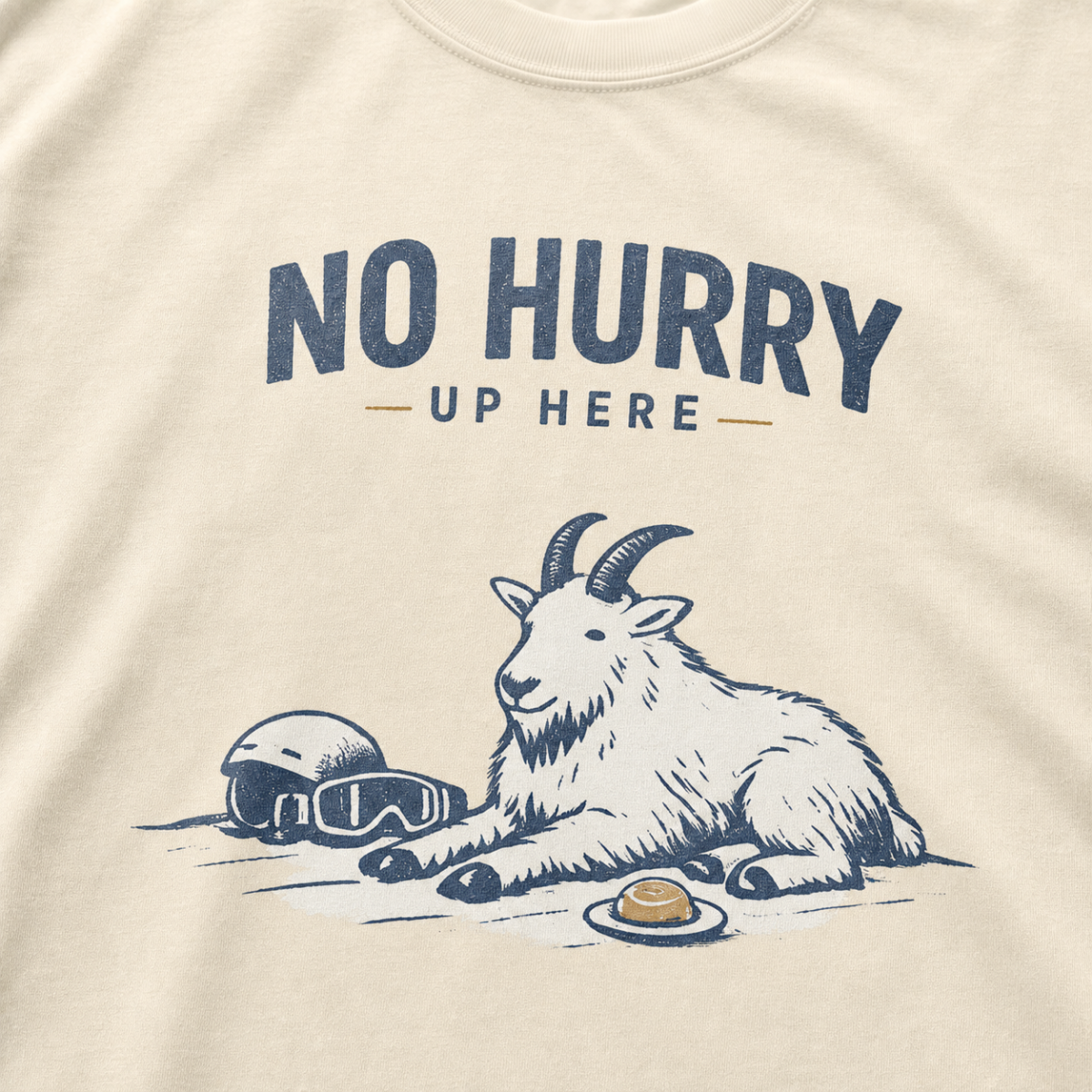 Close-up of an ivory white t-shirt featuring a mountain goat resting in the snow beside a helmet, goggles and snack beneath the words “No Hurry Up Here.”