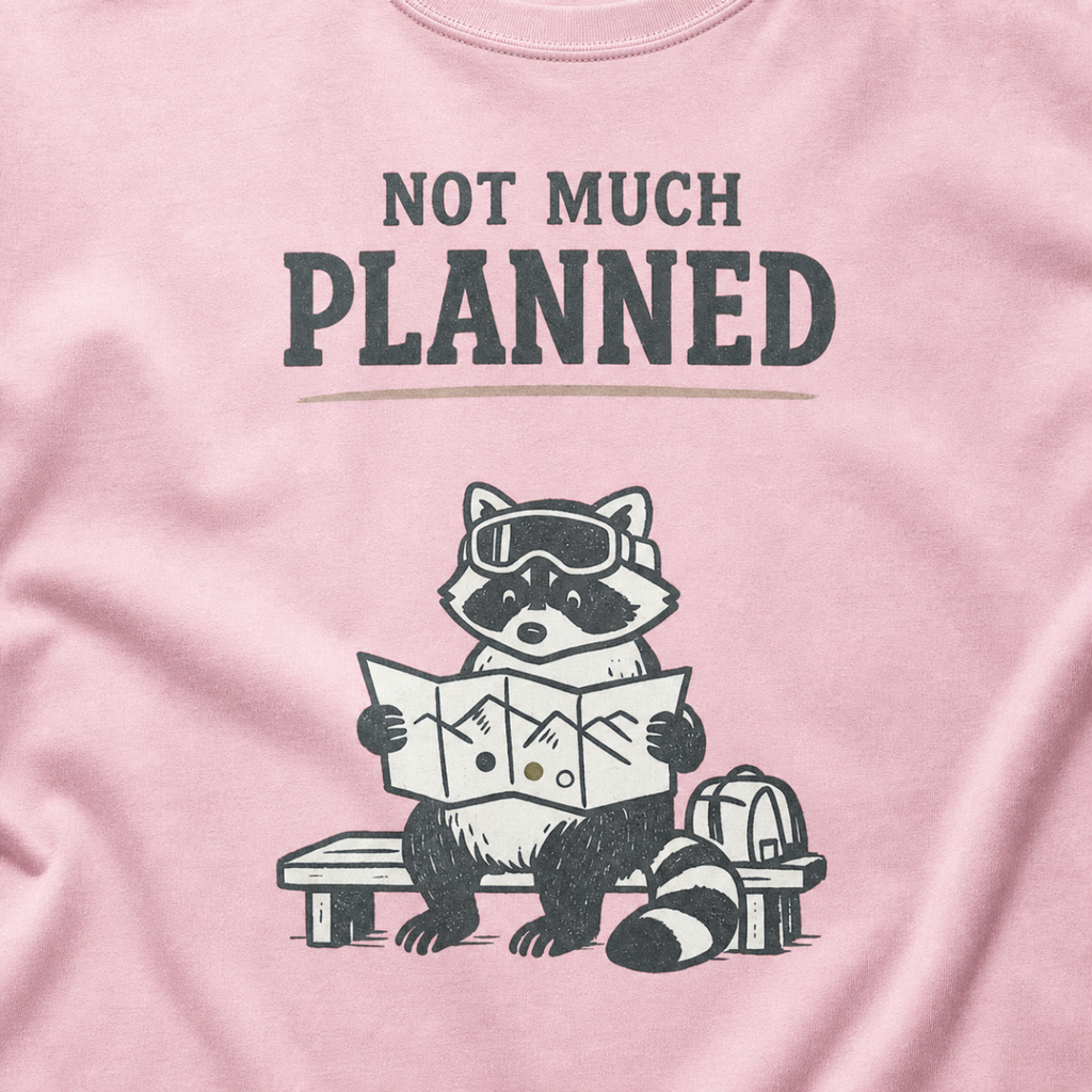 Close-up of a blossom pink t-shirt featuring a raccoon in ski goggles holding a mountain map beside a backpack, with the words “Not Much Planned.”