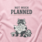 Close-up of a blossom pink t-shirt featuring a raccoon in ski goggles holding a mountain map beside a backpack, with the words “Not Much Planned.”
