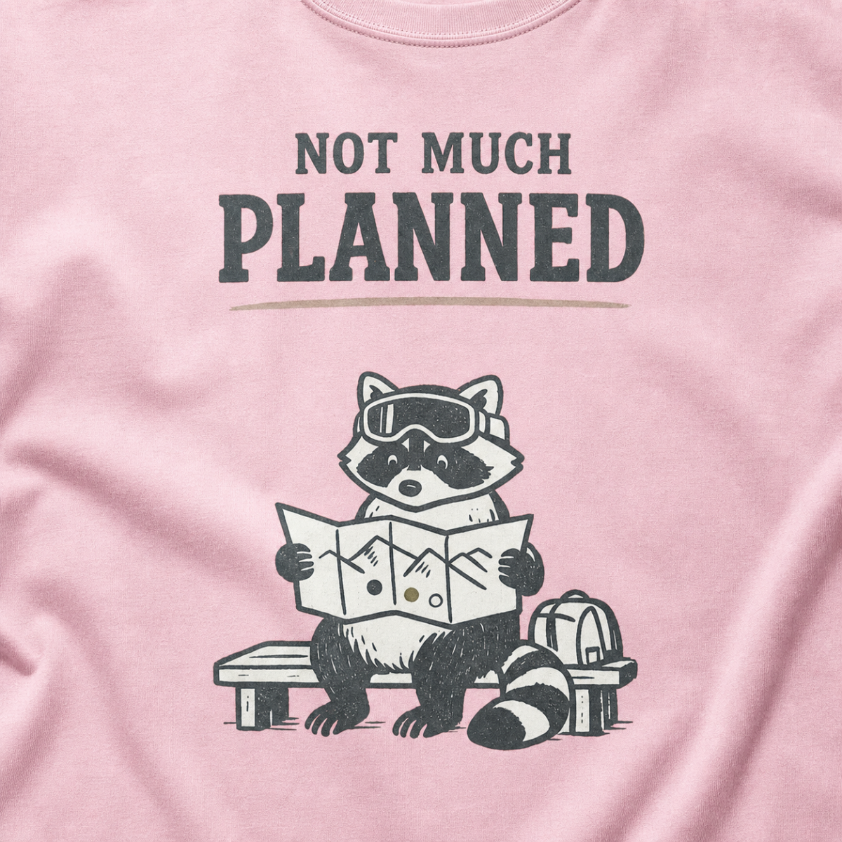 Close-up of a blossom pink t-shirt featuring a raccoon in ski goggles holding a mountain map beside a backpack, with the words “Not Much Planned.”