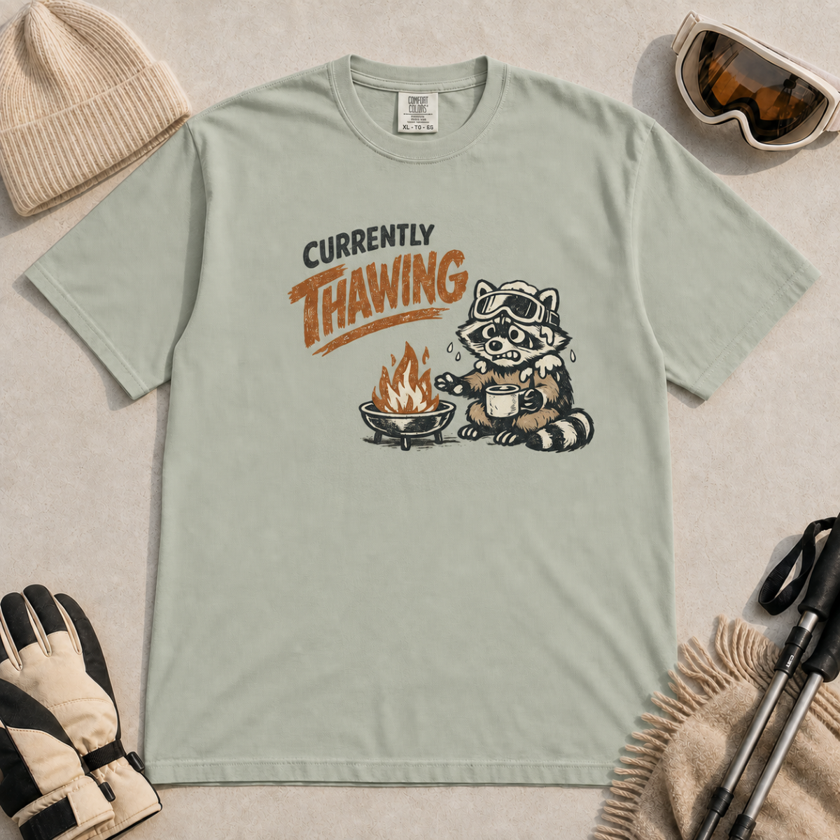 Bay green t-shirt featuring a raccoon warming beside a fire with a mug and the words “Currently Thawing.”