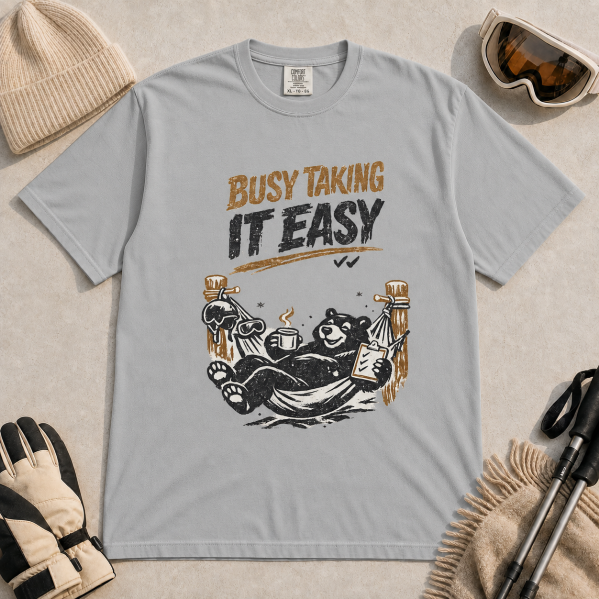Granite grey t-shirt featuring a black bear resting in a hammock with ski gear and the words “Busy Taking It Easy.”