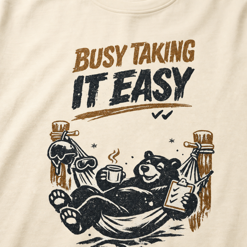 Close-up of a black bear relaxing in a ski-lodge hammock with a hot drink and checklist beneath the words “Busy Taking It Easy.”