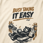 Close-up of a black bear relaxing in a ski-lodge hammock with a hot drink and checklist beneath the words “Busy Taking It Easy.”