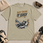 Khaki t-shirt featuring a black bear resting in a hammock with ski gear and the words “Busy Taking It Easy.”