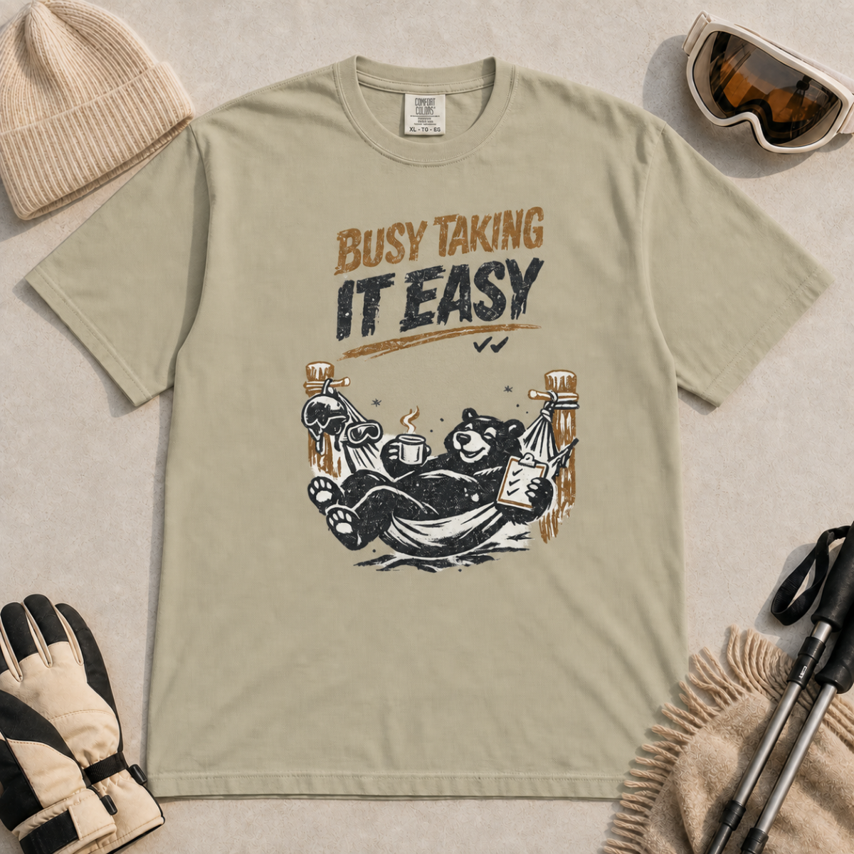 Khaki t-shirt featuring a black bear resting in a hammock with ski gear and the words “Busy Taking It Easy.”