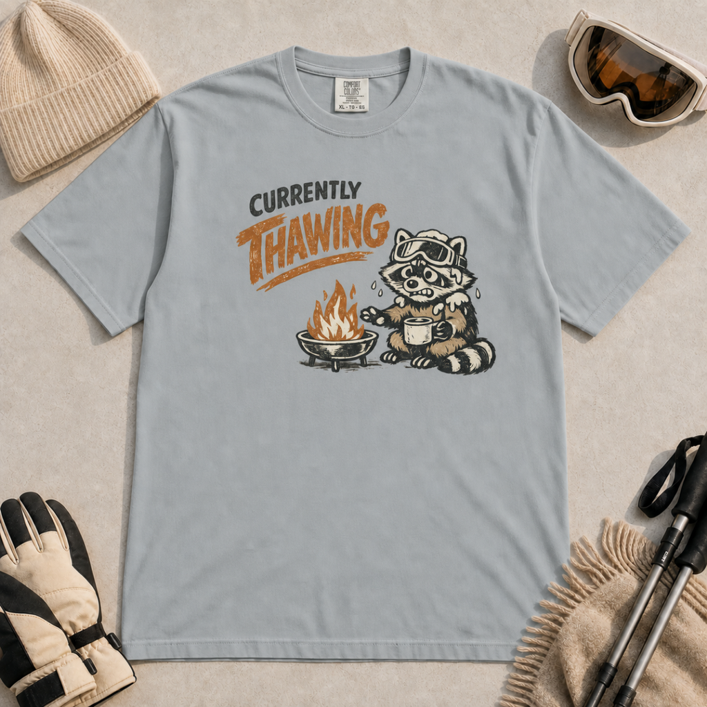 Granite gray t-shirt featuring a raccoon warming beside a fire with a mug and the words “Currently Thawing.”