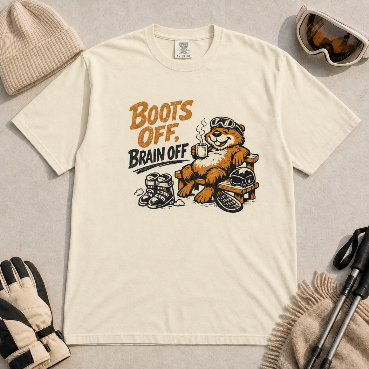 Ivory white t-shirt featuring a marmot relaxing on a bench with ski gear and the words “Boots Off, Brain Off.”