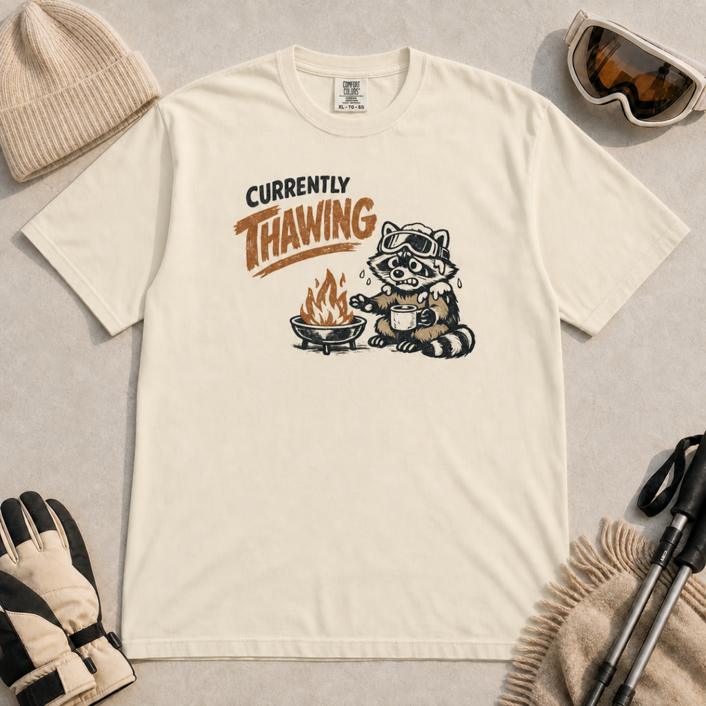 Ivory white t-shirt featuring a raccoon warming beside a fire with a mug and the words “Currently Thawing.”