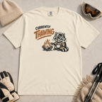 Ivory white t-shirt featuring a raccoon warming beside a fire with a mug and the words “Currently Thawing.”