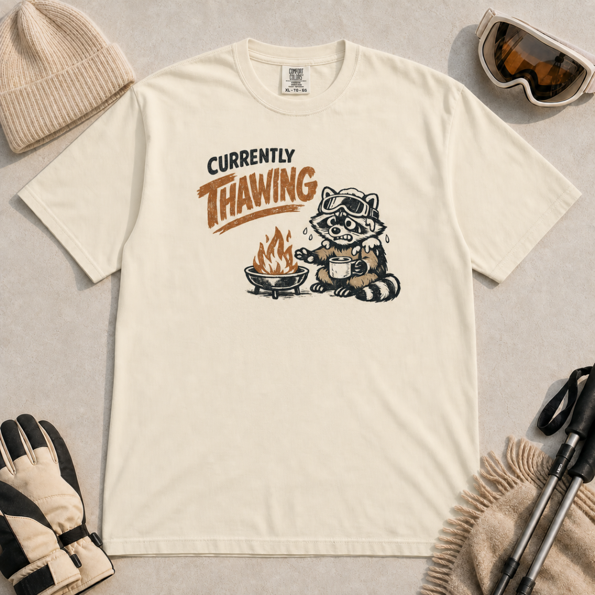 Ivory white t-shirt featuring a raccoon warming beside a fire with a mug and the words “Currently Thawing.”