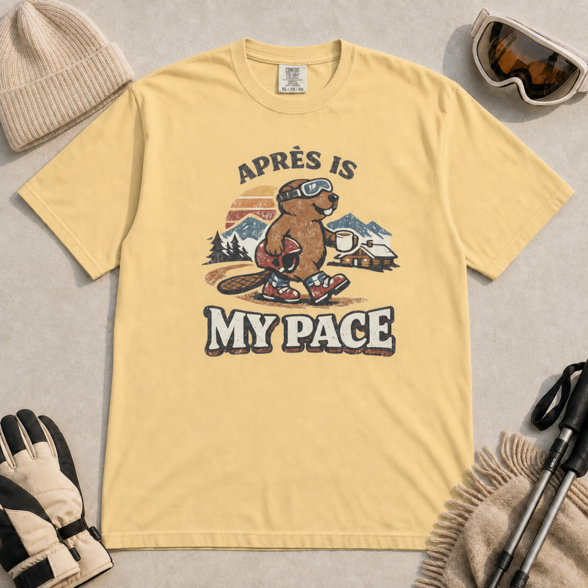 Butter yellow t-shirt featuring a beaver in ski boots holding a mug beneath the words “Après Is My Pace.”