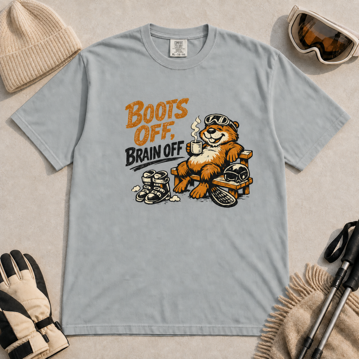 Granite gray t-shirt featuring a marmot relaxing on a bench with ski gear and the words “Boots Off, Brain Off.”