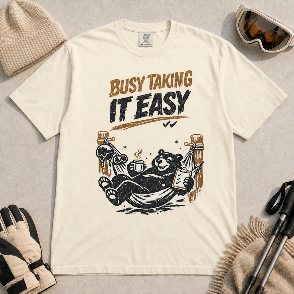 Ivory white t-shirt featuring a black bear resting in a hammock with ski gear and the words “Busy Taking It Easy.”
